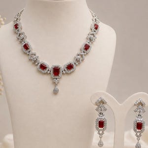 May include: A silver-toned necklace and matching drop earring set featuring rectangular red stones surrounded by small clear crystals. The necklace has a central pendant with a small teardrop-shaped accent, and the earrings mirror this design.