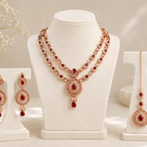 May include: A jewelry set featuring a double-strand necklace, two drop earrings, and a maang tikka. Each piece is crafted with rose gold-toned metal, clear crystals, and prominent teardrop-shaped red stones arranged in intricate floral patterns.