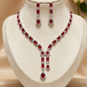 May include: A jewelry set consisting of a necklace and matching drop earrings. The necklace features a series of rectangular red stones linked by small clear stones, with a central drop design. The earrings mirror this red and clear stone pattern.