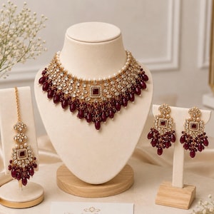 May include: A gold-toned jewellery set featuring a wide choker necklace, a pair of drop earrings, and a maang tikka. Each piece is adorned with square-cut deep red stones, clear white crystals, and dangling teardrop-shaped dark red beads.