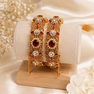 May include: Two gold-toned metal bangles featuring intricate filigree patterns, embedded with oval-shaped deep red stones and small clear white crystals. Each bangle includes a hinged opening with a small attached safety chain for secure closure.