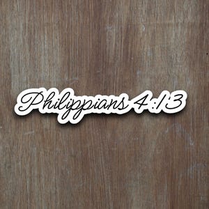 May include: A white sticker with the text "Philippians 4:13" in a cursive font with a black outline. The sticker is placed on a wooden surface with a brown color and wood grain.