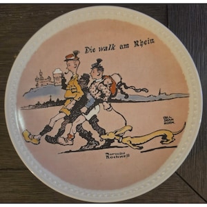 May include: A round ceramic decorative plate with a light pink background featuring a Norman Rockwell illustration titled Die walk am Rhein. The image depicts two figures walking with a dog, a castle in the distance, and the text Bill Dean Norm.