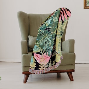 Op de afbeelding: Een groene fauteuil met een deken met een tropisch bladmotief in tinten groen, roze en zwart. De deken heeft een gefranst randje.