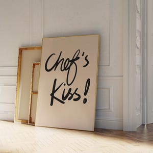 May include: A white canvas with black lettering that reads "Chef's Kiss!" The canvas is leaning against a white wall and a wooden floor.
