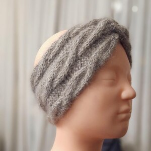 Puede incluir: Una diadema gris tejida con un diseño acanalado texturizado. La diadema está hecha de un material suave, posiblemente lana. El accesorio se muestra en una cabeza de maniquí, destacando su forma y ajuste. El color neutro lo hace versátil.