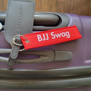 Op de afbeelding: Een paarse koffer met een rood bagagelabel waarop in witte letters "BJJ Swag" staat. De koffer heeft een zwarte handgreep en een zwarte rits.
