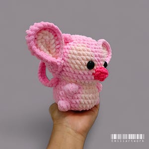 May include: A handmade pink and white crocheted mouse toy with large ears, black eyes, and a red nose. The plush toy is held in a hand against a grey background. The toy is made of soft, textured yarn.