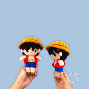 Könnte beinhalten: Zwei handgefertigte Häkelpuppen einer Figur mit gelbem Strohhut, roter Weste, blauen Shorts und schwarzen Haaren. Die Puppen werden vor einem hellblauen Hintergrund gehalten. Die Puppen sind etwa 13 cm groß.