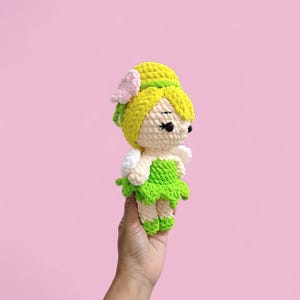 以下が含まれることがあります： 手作りのかぎ針編みの妖精人形。黄色い帽子とピンクの花、緑のドレス、白い羽が特徴です。人形はピンク色の背景に保持されています。