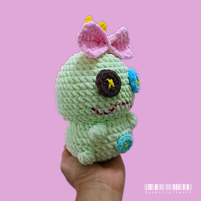 May include: A handmade, light green crochet doll with a pink bow, yellow accents, and button eyes. The doll has a stitched mouth and is held in a hand against a pink background. The doll is a unique, handcrafted item.