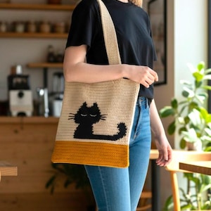 Puede incluir: Bolso tote de crochet beige con un diseño de gato negro. El bolso tiene un borde inferior amarillo mostaza y una correa a juego. El gato está representado con los ojos cerrados y una cola curvada.