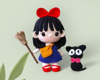 Puede incluir: Una muñeca de crochet con cabello negro, un lazo rojo y un vestido azul sostiene una escoba marrón. La muñeca está de pie junto a un gato negro de crochet con un lazo rosa.