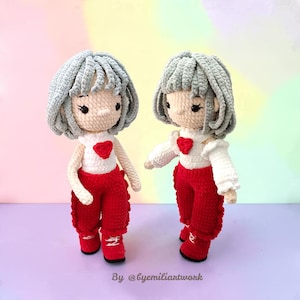 May include: Two handmade crochet dolls with gray yarn hair, white tops featuring a red heart appliqué, and red pants. The dolls have black embroidered eyes and are standing upright. The text By @byemiliartwork is visible at the bottom.