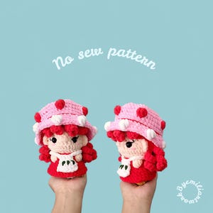 May include: Two handmade crochet dolls, each held in a hand, feature a red dress, red curly hair, and a pink hat with red and white accents. The dolls have white aprons and are set against a light blue background. The text "No sew pattern" is visible.