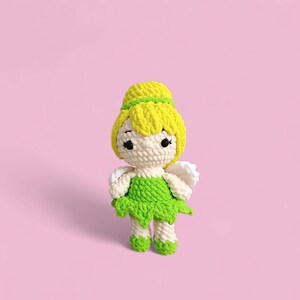 以下が含まれることがあります： 黄色い髪、緑色のドレス、白い羽を持つ手作りのかぎ針編み人形。人形は黒い目を持ち、ピンク色の背景に立っています。人形の高さは約15cmです。