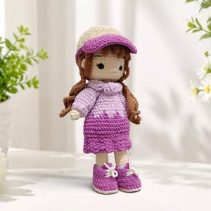 May include: A handmade crochet doll featuring brown yarn hair, a cream and purple baseball cap, and a purple gradient sweater dress. The doll has embroidered facial features and wears matching purple crochet sneakers with white laces.