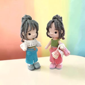 May include: Two handmade crochet dolls with dark gray yarn hair styled in buns. The left doll wears a white top and blue skirt. The right doll wears a green top, pink pants, and carries a small white and pink crocheted bag. Both have simple facial features.