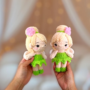 May include: Two handmade crochet dolls, each featuring blonde hair, a green skirt, and a pink flower accent. The dolls have delicate facial features and are held up by hands. The background is blurred, with warm tones.