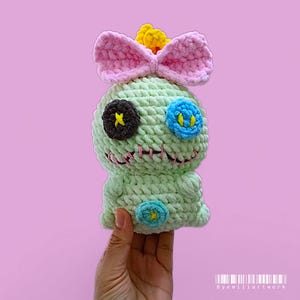 May include: A handmade, light green crochet doll with a pink bow, yellow crown, and stitched facial features. The doll has a dark brown eye with a yellow cross, a blue eye with yellow details, and a blue button on the body. The doll is held against a pink background.