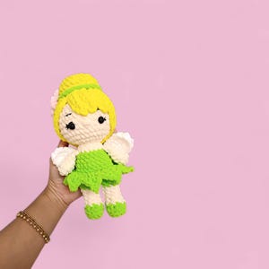 以下が含まれることがあります： 黄色い髪、緑のドレス、白い羽を持つ手作りのかぎ針編みの人形。人形は黒い目を持ち、ピンクの背景に保持されています。人形の髪は、緑のヘアバンドとピンクの花でまとめられています。