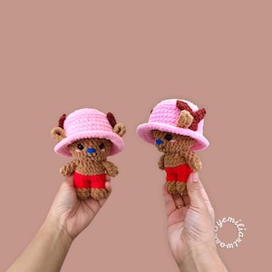 May include: Two handmade crochet figures of a cartoon character, each wearing a pink hat and red shorts. The figures have brown bodies, blue noses, and brown antlers. The background is a solid, muted pink.