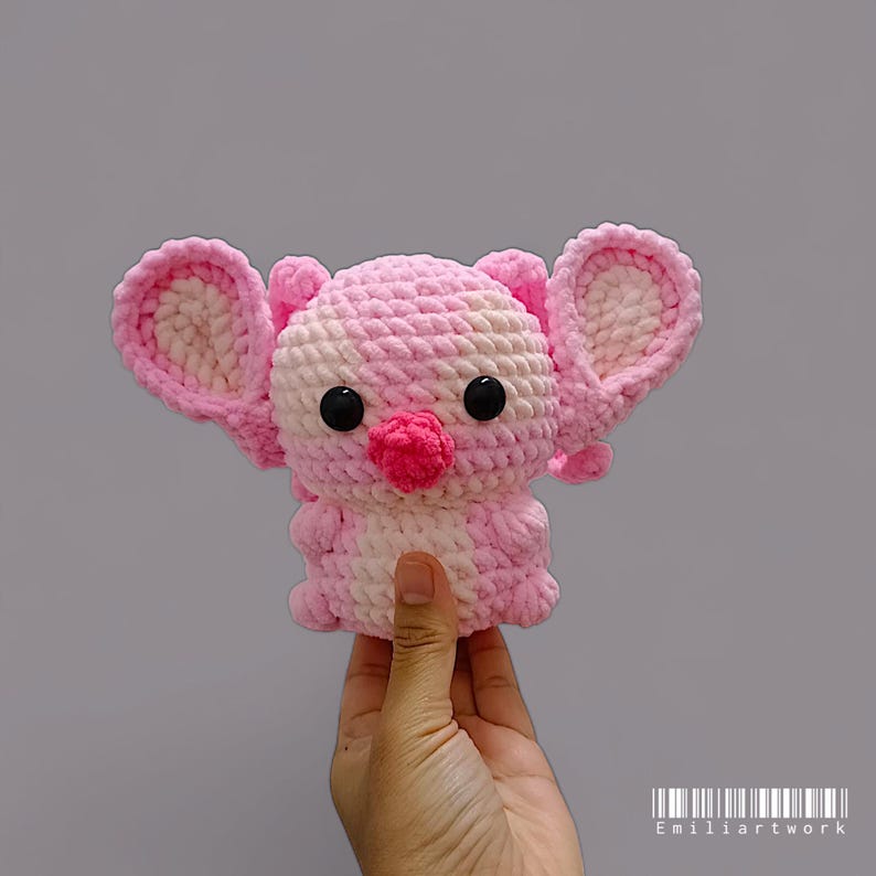May include: A handmade pink and white crochet animal with large ears, black eyes, and a small pink nose. The plush toy is held in a hand against a grey background. The toy is a cute, soft, and cuddly creation.