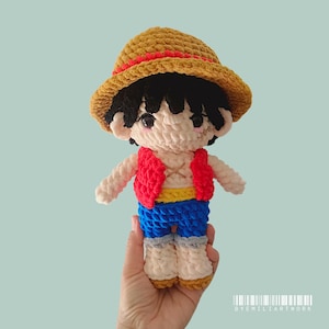 May include: A handmade crochet doll of a character wearing a straw hat with a red band, a red vest, blue shorts, and yellow belt. The doll has black hair and is held in a hand. The doll is approximately 18 cm tall.