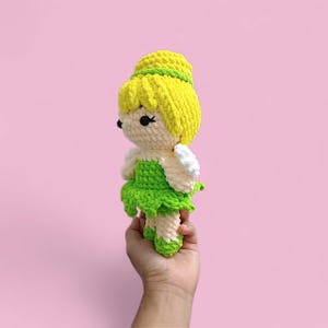 以下が含まれることがあります： 黄色い髪、緑のドレス、白い羽を持つ手作りの編み人形。人形はピンク色の背景に対して手で持たれています。妖精は黒い目と緑色のヘアバンドをしています。