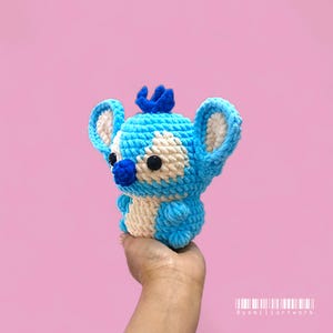 May include: A blue and white crochet toy, featuring large ears and a crown. The handmade plush has black button eyes and a blue nose. The toy is held in a hand against a pink backdrop.