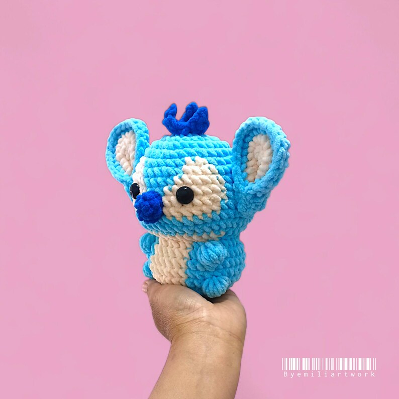 May include: A blue and white crochet toy, featuring large ears and a crown. The handmade plush has black button eyes and a blue nose. The toy is held in a hand against a pink backdrop.