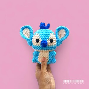 May include: A handmade blue and white crochet Stitch doll with large ears, a small nose, and black eyes. The doll is held against a pink background. The doll is a character from the animated movie Lilo & Stitch.