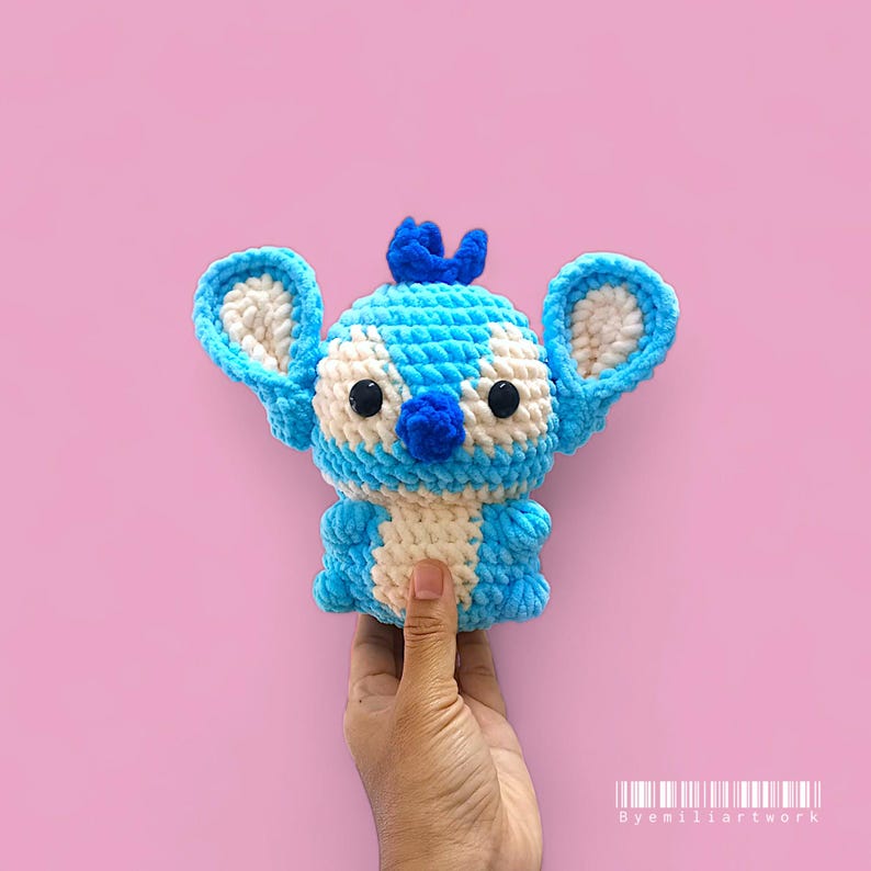 May include: A handmade blue and white crochet Stitch doll with large ears, a small nose, and black eyes. The doll is held against a pink background. The doll is a character from the animated movie Lilo & Stitch.