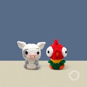 May include: Two handmade crochet figures: a white pig with gray accents and a red bird with large white eyes. The pig has a pink snout and the bird has a green and yellow body. Both are on a light tan surface against a blue background.