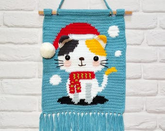 Puede incluir: Un tapiz de pared de ganchillo hecho a mano con un gato calicó que lleva un gorro y una bufanda de Papá Noel. El gato está sobre un fondo azul con detalles de copos de nieve blancos y una borla. El tapiz de pared está suspendido de una clavija de madera.
