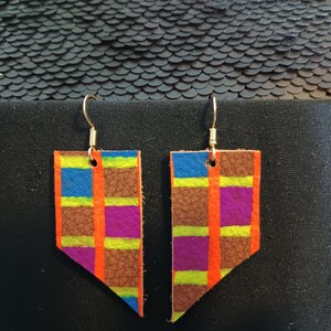 Könnte beinhalten: Ein Paar Lederohrringe mit geometrischem Muster. Die Ohrringe sind braun mit orangefarbenen, violetten, grünen und blauen Streifen.