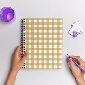 Może przedstawiać: Szyty spiralnie notes z brązowo-białą kratką na okładce. Notes leży płasko na białym tle z fioletowym długopisem i fioletowym spinaczem.