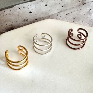 May include: Three minimalist rings in gold, silver, and copper. Each ring is made of three thin bands of metal that are twisted together.