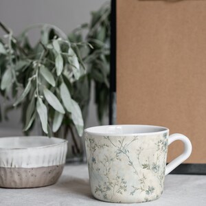 May include: A white ceramic mug with a floral pattern in shades of blue and green. The mug has a handle and is sitting on a white tablecloth.