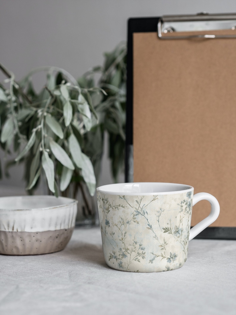 May include: A white ceramic mug with a floral pattern in shades of blue and green. The mug has a handle and is sitting on a white tablecloth.
