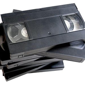 May include: A stack of black VHS video cassette tapes. Each tape has a rectangular shape with a textured surface and clear plastic windows revealing the internal spools. The tapes are arranged in a slightly haphazard pile.