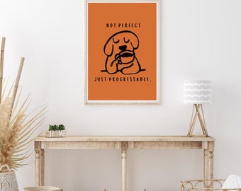 May include: A framed art print with an orange background features a black line drawing of a dog holding a coffee cup. The text above reads "NOT PERFECT" and below "JUST PROGRESSABLE."