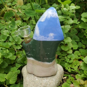 May include: A ceramic pipe shaped like a mountain with a blue sky and white clouds on top, a green forest in the middle, and a sandy beach at the bottom. The pipe has a glass bowl attached to the side.
