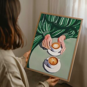 May include: A painting of two cups of coffee on a green and white striped tablecloth. The cups are white with brown coffee and a layer of foam on top. The painting is framed in a light wood frame.