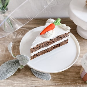 Puede incluir: Una rebanada de pastel de zanahoria con glaseado blanco y una decoración de zanahoria en un plato blanco. El pastel tiene tres capas de bizcocho marrón oscuro y glaseado blanco. La zanahoria es naranja con hojas verdes.