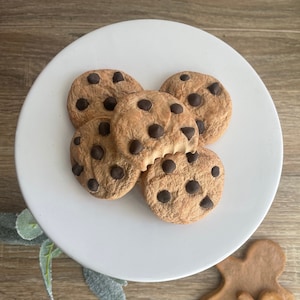 Pode incluir: Um suporte branco para bolos com cinco biscoitos com pepitas de chocolate, um com uma mordida. Pepitas escuras salpicam os biscoitos. Dois biscoitos de gengibre estão ao lado.