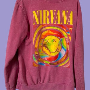 May include: A pink sweatshirt featuring the word "NIRVANA" in yellow block letters above a colorful smiley face design. The graphic includes a yellow smiley face with crossed-out eyes and a tongue sticking out, surrounded by rainbow-colored swirls.