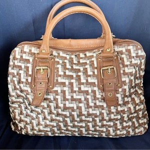 May include: A woven leather handbag with brown and white straps and a brown leather handle. The bag has two buckles on the front and a zipper closure.