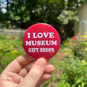 以下が含まれることがあります： 「I LOVE MUSEUM GIFT SHOPS」の白い文字が入った赤い丸いボタン。ボタンは手で持たれており、背景にはぼやけた緑色の木々や建物があります。