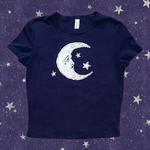 Può includere: T-shirt blu navy con un motivo a mezzaluna bianca con un volto e stelle. La grafica è stampata sulla parte anteriore della maglietta a maniche corte. La maglietta è in tessuto morbido.